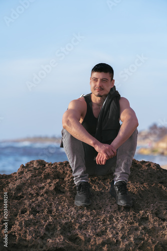 A young man of athletic build, with short dark hair, a dark beard, and mustache, sits on a large porous rock on the shore of the Mediterranean Sea.