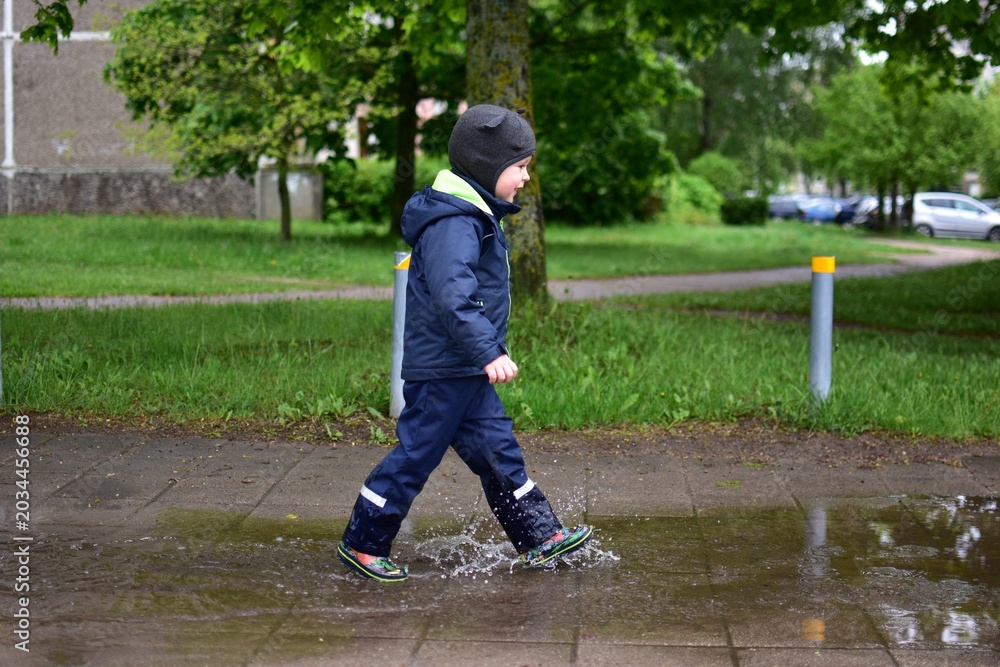 Fototapeta premium Toddler walking through rain puddles in waterproof outdoor clothing on a wet city sidewalk after spring rain. Playful childhood moment with splashing water and urban greenery in background.