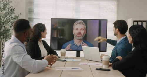 Multicultural business team greets remote manager on video call during online meeting in office. Coworkers wave at screen and welcome speaker on conference