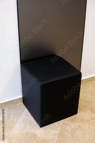 Black leather cube ottoman stool next to dark panel in modern dental clinic interior. Waiting area furniture concept