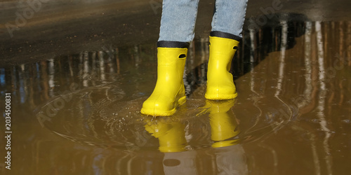 Bild auf Leinwand Female legs walking through spring puddle in yellow waterproof boots creating wa