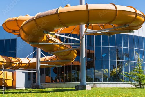 Large yellow water slide spiral tube outside an indoor aquapark building with glass windows on a bright sunny summer day