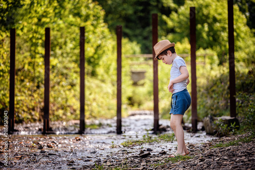 Thoughtful boy standing barefoot by shallow forest river looking down