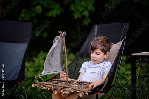 Young boy relaxing in camping chair holding DIY wooden stick boat toy