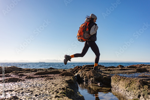 Active female hiker with orange backpack jumping over rocky coastal terrain at low tide, dynamic seaside adventure scene with motion, balance, freedom, ocean horizon, and copy space sky