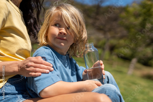 Cuadro en lienzo Little blond boy holding water bottle sitting with mother on grass in sunny park