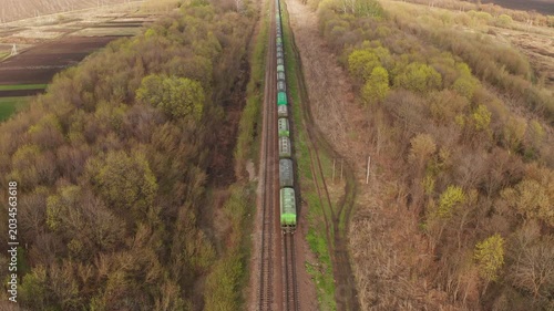 Aerial video of long freight train with green grey cars moving on railway tracks through a spring forest. Drone follows train in rural countryside with fields trees. Logistics transport concept
