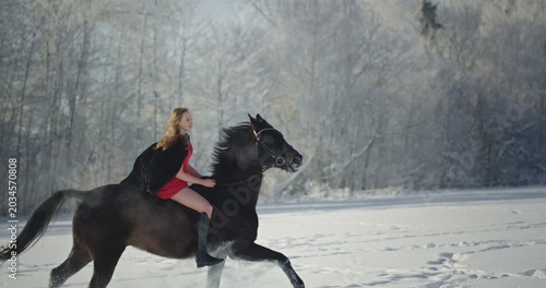 Young woman riding black horse galloping in winter snow