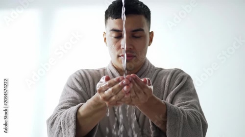 Serene young adult, neutral garment, splashes pristine water on face in bright minimalist spa, slow-motion droplets glisten, bokeh. Concept of refreshing rejuvenation and well-being