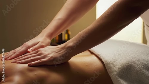 Smooth tracking video of gender-neutral hands performing a back and shoulder massage in a luxurious spa with warm light, white towels, bokeh. Concept of ultimate relaxation