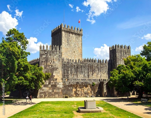 A stone fortress stands prominently beneath a bright blue sky, flanked by verdant trees, walkways, and a well-kept lawn