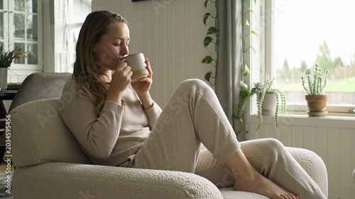 A person in neutral loungewear sips tea or reads in a cream bouclé armchair within a serene scandinavian home, bathed in soft daylight. Hygge lifestyle concept