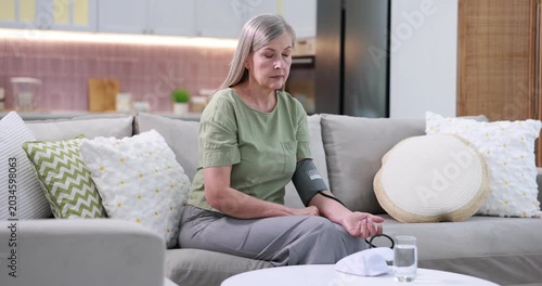 Senior woman measuring blood pressure on sofa at white table indoors. Camera moving forward