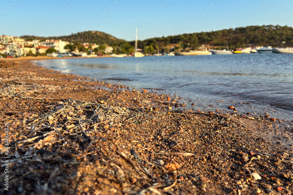 Fototapeta premium Scenic Turkish Aegean coast with pebble beach, calm blue sea, and boats moored near tranquil seaside resort town on a sunny day; ideal for summer travel, Mediterranean vacation, and tourism concept