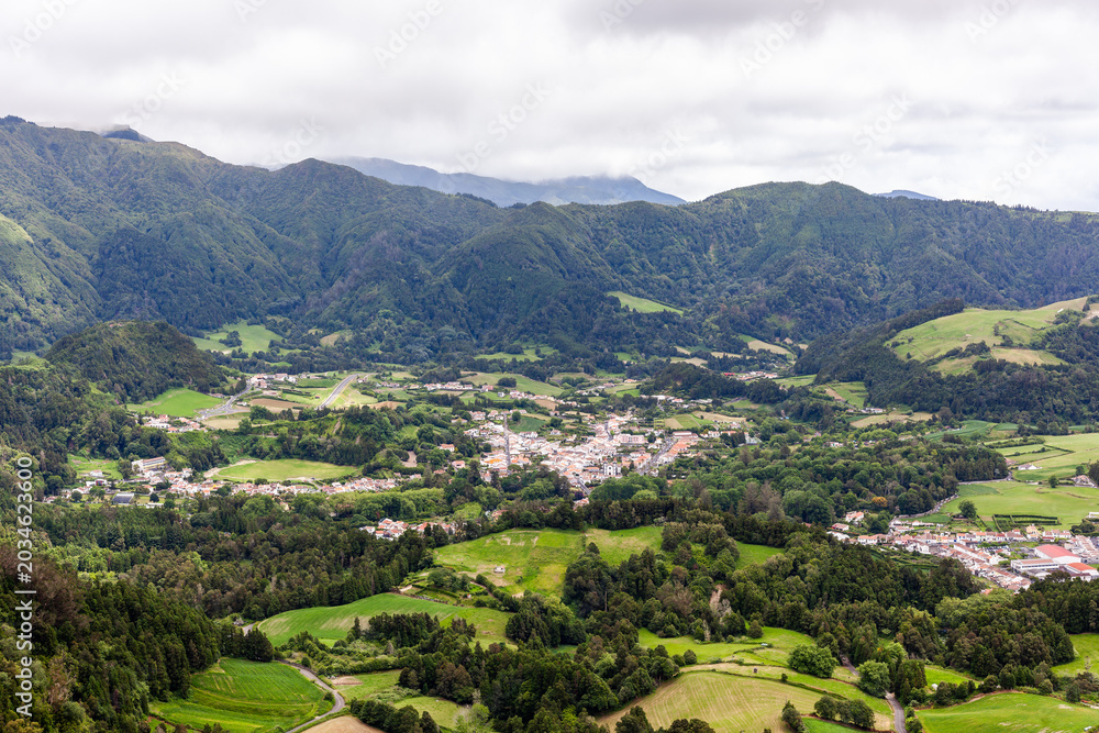 Fototapeta premium Furnas village nestled in lush green valley with surrounding mountains. Scenic view from Pico do Ferro Viewpoint showcasing volcanic landscape.