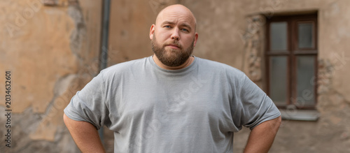 Portrait of obese man standing outdoors near wall, neutral expression, bald head, beard, gray shirt, front view