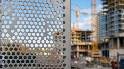 Perforated metal sheet fence in front of a construction site with cranes and unfinished highrise buildings in the background