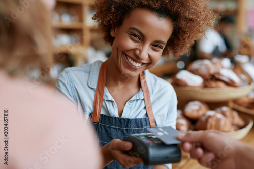 Warm smile meets customer at cozy bakery counter. Vendor wears denim apron, accepts card payment. Soft-focus background holds pastries, wood shelves. Customer hands card toward portable payment device