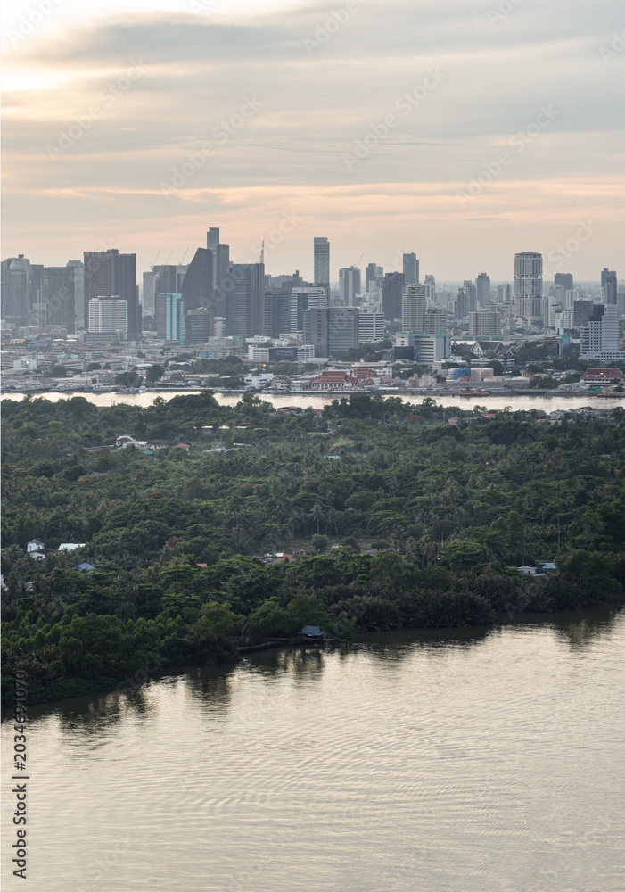 Fototapeta premium A vast expanse of dense, emerald canopy blankets the peninsula of Bang Kachao, standing as a tranquil fortress of nature against the encroaching modern world. In the foreground, the glassy waters of t