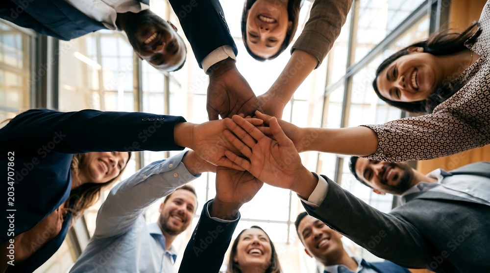 Obraz premium Group of diverse professionals gather in a high-ceilinged office space. They form a unified hand stack, smiling broadly at the camera. Their joyful expressions convey teamwork and shared success