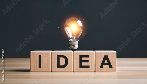 Glowing light bulb above wooden blocks spelling IDEA on dark background