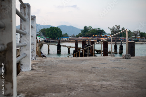 Fotografie Concrete harbor ramp with metal railings leading toward calm tropical water, featuring weathered mooring posts and a lone pigeon near a quiet coastal village in Thailand