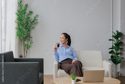 Schilderij op canvas Asian businesswoman enjoying a coffee break, sitting on a sofa in a modern offic