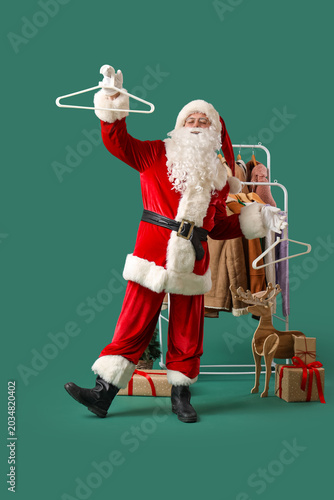 Santa Claus holding hangers near rack with different clothes on green background
