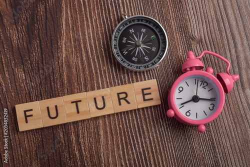 Wooden letter blocks spelling future with compass and pink alarm clock representing time management, planning, future goals, direction, scheduling, and personal development.