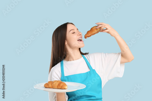 Pretty young woman in apron with fresh croissant on blue background