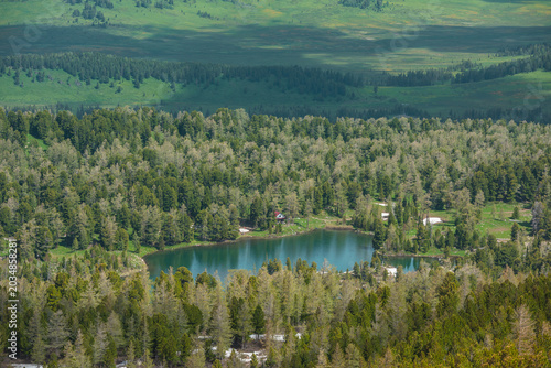 Scenic aerial top view to wood lake against woody hills. Colorful view with cabin on shore of forest lake. Sunlight and shadows of clouds on hilly vastness. Coniferous trees reflection in alpine lake.