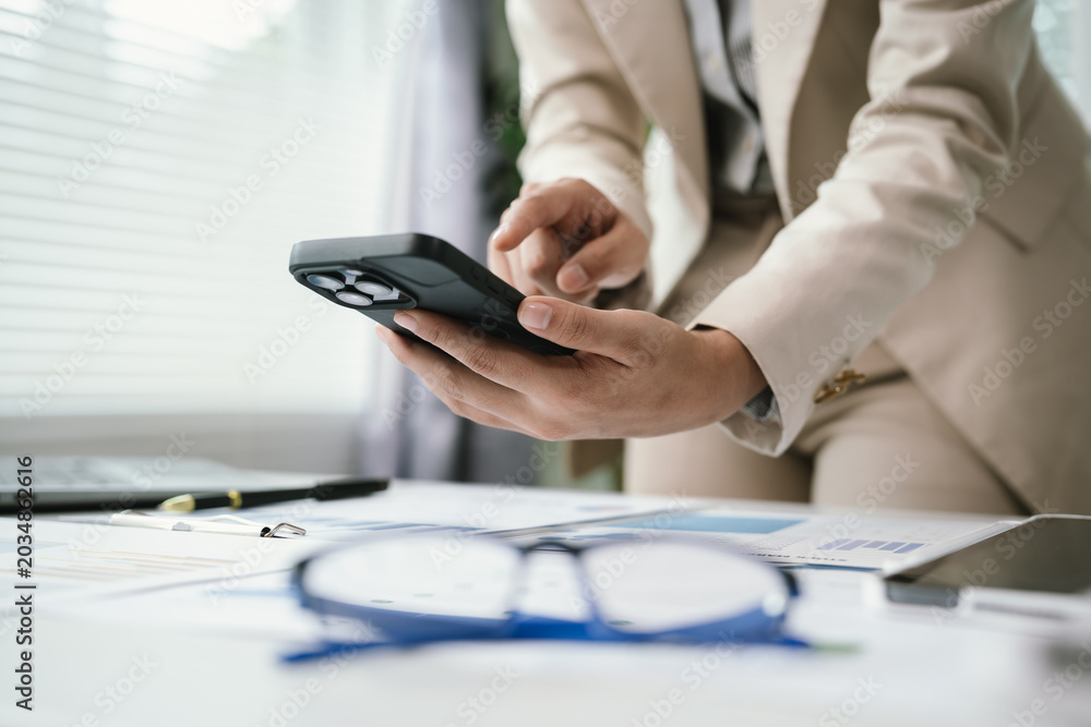 Fototapeta premium Close-up of a business professional using a smartphone while reviewing financial documents at a modern office desk, highlighting mobile communication, productivity, and digital workflow.