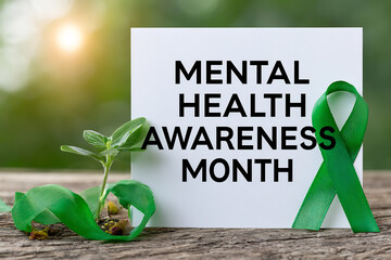 A white card with 'MENTAL HEALTH AWARENESS MONTH' text, a green ribbon, and a small plant sprout on a wooden surface with a blurred green background and sunlight.