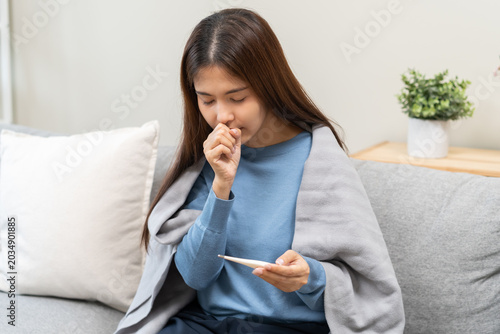 Sick, influenza asian young woman, girl have fever, cough and sore throat, holding thermometer for check measure body temperature, illness while sitting under blanket rest on sofa at home. Health care