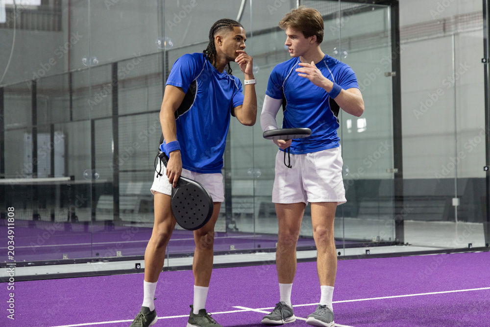 Obraz premium Diverse male partners talking and gesturing on purple court in blue shirts holding paddles