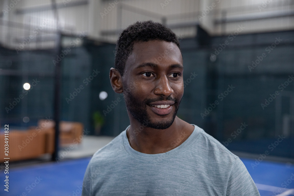 Fototapeta premium Smiling African-American man in gray tee standing on court with blue floor, mesh fence, brown couch