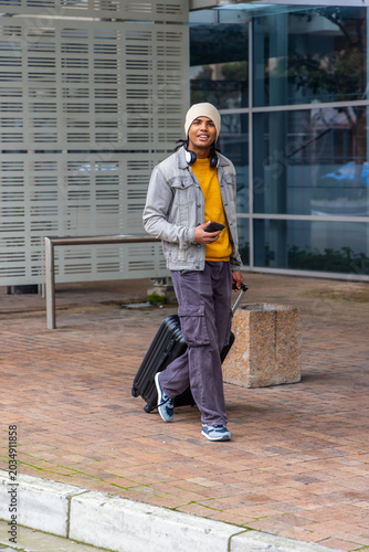 Man walking across brick plaza pulling black suitcase and holding smartphone wearing denim jacket