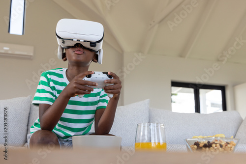 Fotografía African American male child sitting at home wearing VR headset, holding controll