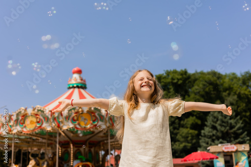 happy little girl is blowing bubbles in front of a carousel at an amusement park.