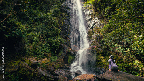 Fotografie Woman sitting on rock contemplating tall waterfall in lush tropical rainforest E
