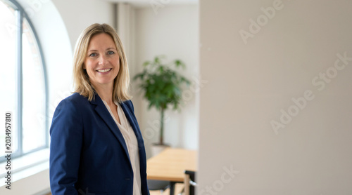 A confident blonde businesswoman in a blue suit smiles warmly in a bright modern office setting, representing professional success, corporate leadership, and a positive workspace atmosphere.