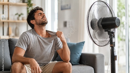 Man Cooling Off With Electric Fan During Summer Heatwave Inside Modern Living Room at Home