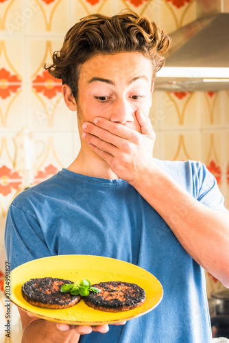 Tableau sur toile Young man in a kitchen reacts with shock while holding a plate of burnt patties,