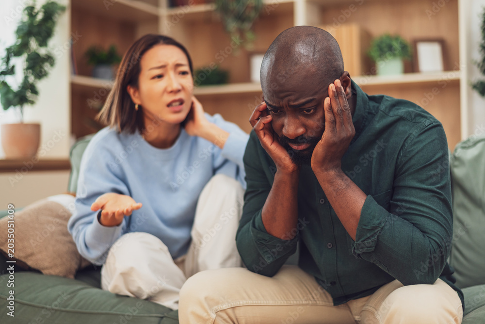 Fototapeta premium Multiracial couple sitting on sofa at home and arguing. Man is angry and woman is apologizing to him. Relationship difficulties.