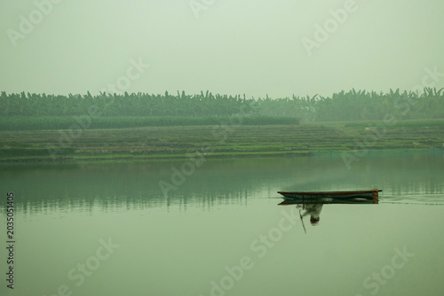 A lone fishing boat rests on a mirror like river as fog drifts