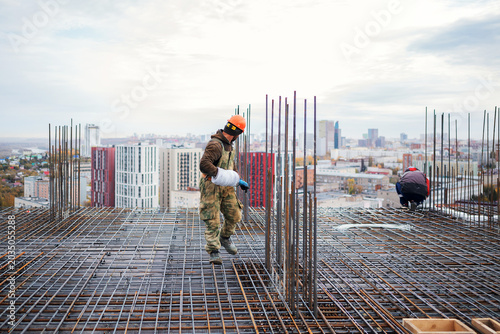 фотографія A construction worker in a hard hat carrying a coil of wire walks across a vast steel rebar grid preparing for concrete pouring on a high-rise building site with a city view