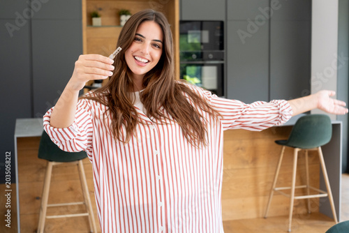 Tableau sur toile Smiling woman holding keys in modern kitchen, celebrating moving into a new home with excitement and pride; casual striped shirt, open arms, bright interior, real estate and lifestyle concept