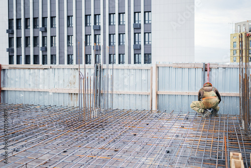 Tableau sur toile An ironworker in a hard hat crouches on a steel rebar grid, tying reinforcement near a corrugated metal safety fence on a high-rise construction site with a modern facade behind