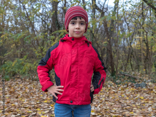 Confident Little Boy in Red Jacket Standing with Hands on Hips in Autumn Forest