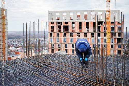 Foto An ironworker bends to tie a steel rebar grid on a high-rise concrete slab, with a brick building under construction and yellow tower cranes in the background